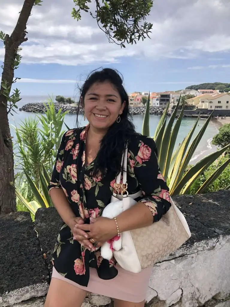 Marcela Arias smiling on vacation near the ocean, wearing a floral top and holding a cream handbag with scenic town and water in the background