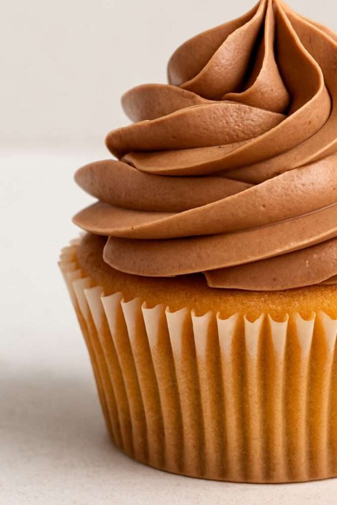 Smooth swirl of fluffy milk chocolate ganache frosting on a vanilla cupcake. Clean neutral background, bakery-style presentation.