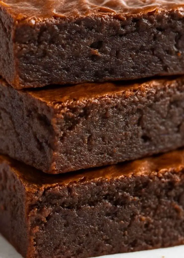 Close-up side view of bakery-style fudgy brownies stacked on parchment paper, showing their dense, moist interior and shiny crackly tops