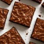Bakery-style fudgy brownies with shiny, crackly tops, sliced into squares on parchment paper to show off their rich chocolate texture and flaky crust.