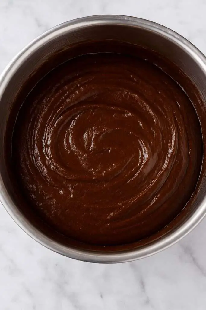 Glossy chocolate brownie batter with a smooth swirl in a stainless steel mixing bowl on a marble background.