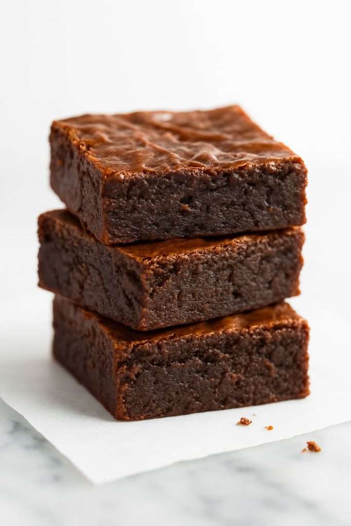 Stack of bakery-style fudgy brownies with shiny, crackly tops and dense, moist chocolate texture shown in a close-up side view.