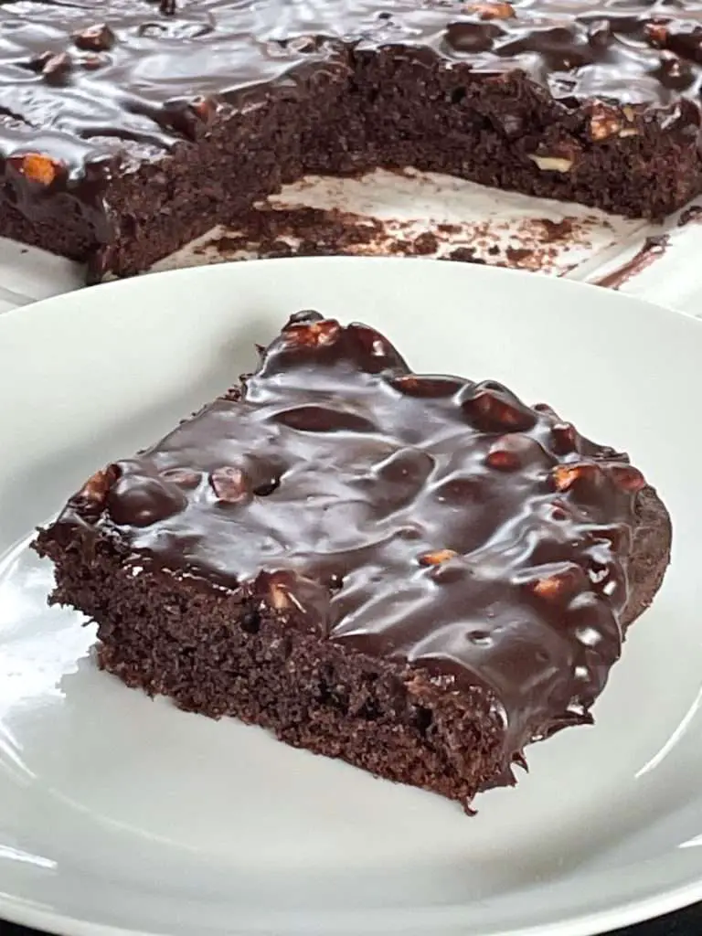 Fudgy Texas sheet cake slice on a white plate with glossy chocolate glaze and chopped pecans, full cake in background.