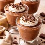 Four glass cups of silky chocolate mousse with custard base, topped with whipped cream and chocolate shavings, ready to serve