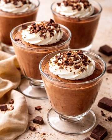Four glass cups of silky chocolate mousse with custard base, topped with whipped cream and chocolate shavings, ready to serve