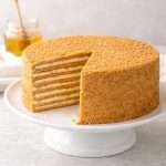 Russian honey cake (Medovik) on a white cake stand with one slice removed showing 8 honey cake layers and creamy filling